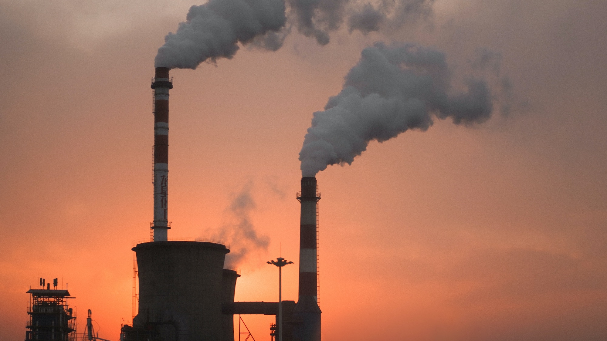 A photo of smoke coming from the chimney of a factory. The sky is orange either due to a wildfire or sunset
