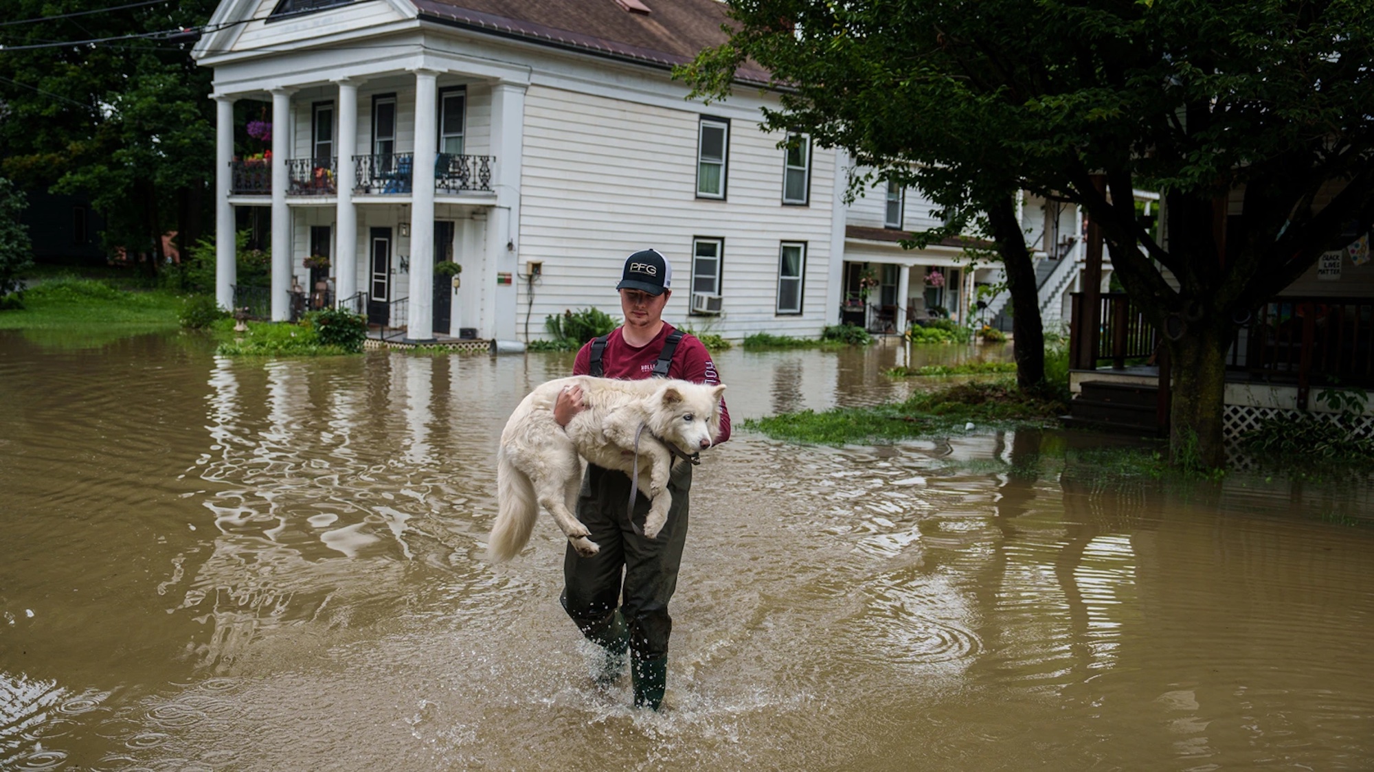 A white man in black pants and a red t-shirt carrying his white big dog through a flooded area