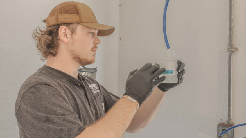 White man with light copper hair and a brown baseball cap measures water in a small plastic cup.