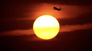 A plane flying in front of a red sky and bright yellow sun