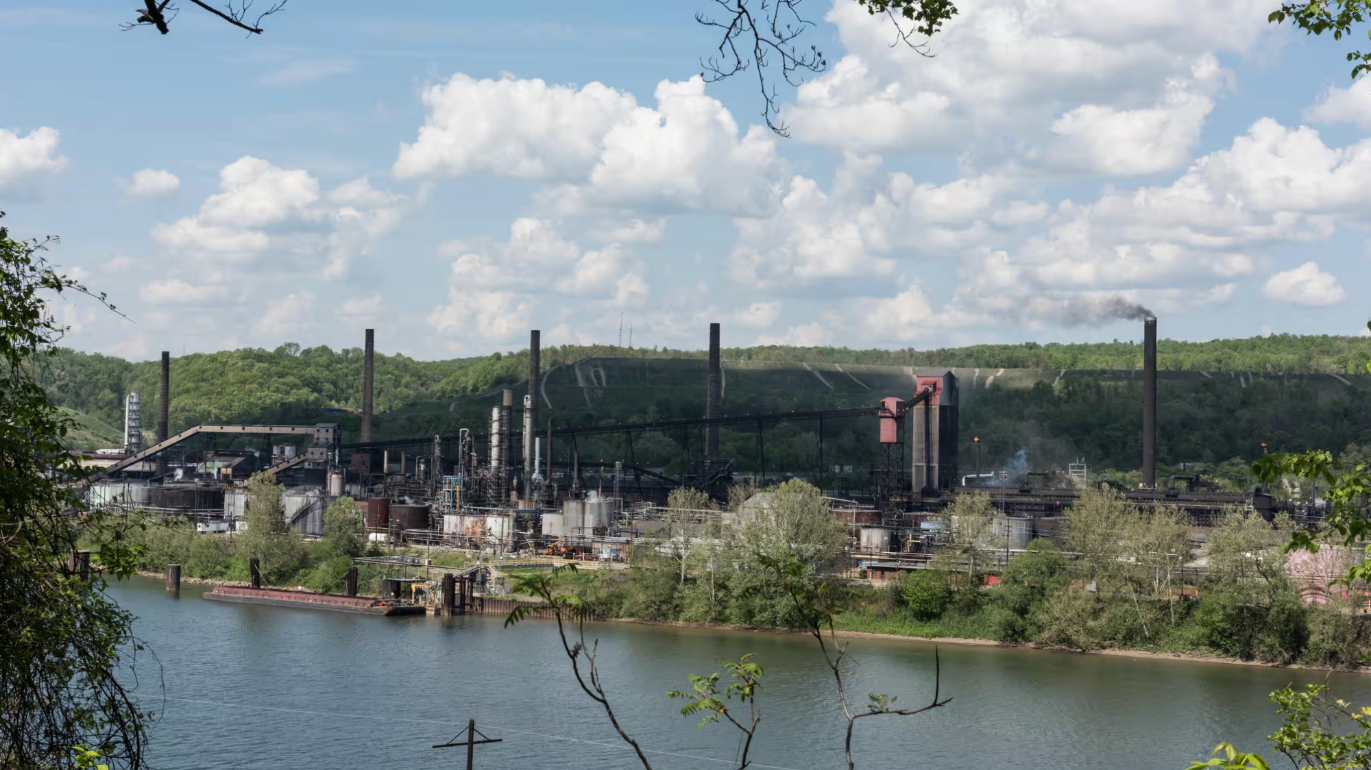 Behind a river, a dark steel plant dominates the mountainside. The light green trees are almost black in its shade and smoke.