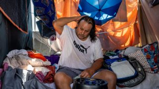 Man with long dark hair sits in a home made of various covered tarps.