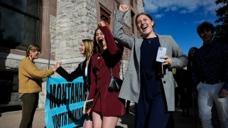 Several youths raise their hands into the air in pride. Behind them, a blue sign partially reads "Montana Youth making"