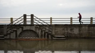 Grey water hits a dark raised walkway, behind it there is a white cloudy sky. A silhouetted person runs across the walkway.