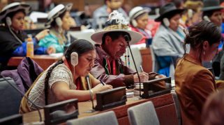 A photo of many people at desks in a large area focuses on two individuals. One has short dark hair and a wide-brimmed white hat. One has long braided dark hair and is wearing a white device on their ear.