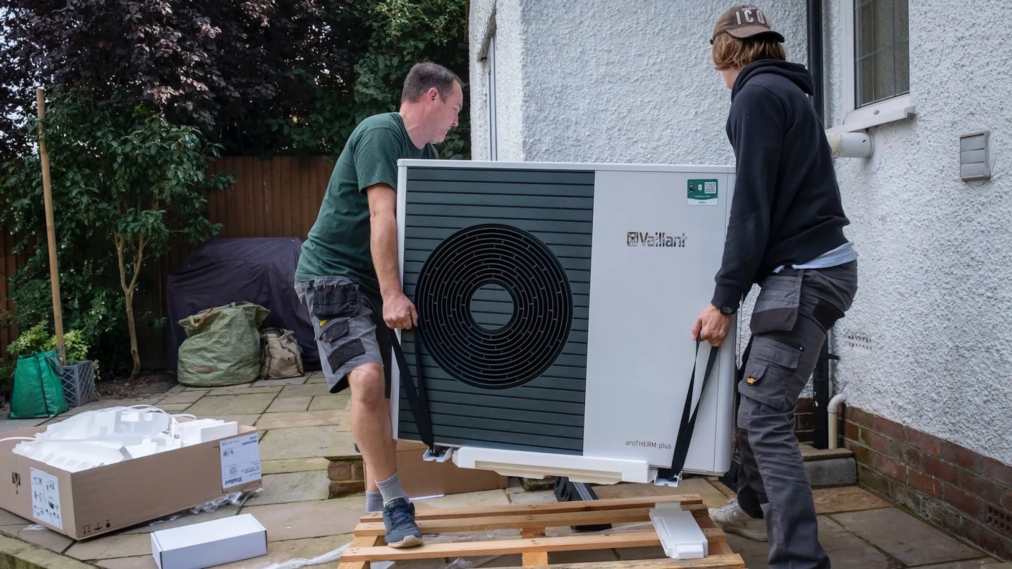 Two white people carrying a heat pump into their home