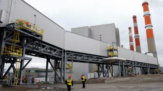 A white building on black stilts sits in front of a white sky. In the background orange towers billow white smoke. In the foreground, two workers stand, small, wearing yellow and orange jackets.
