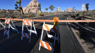 Orange tape and barriers stand in front of a road in front of several rocks and trees.