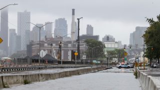 An empty stretch of grey highway is flooded with water. In the background there are cars and streetlights and the New York City skyline. Everything is covered in a rainy mist.
