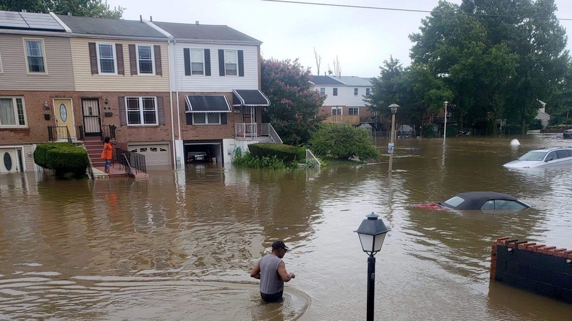 Flooding in Eastwick, showing parts of housing underwater, cars and a Black person walking through the water