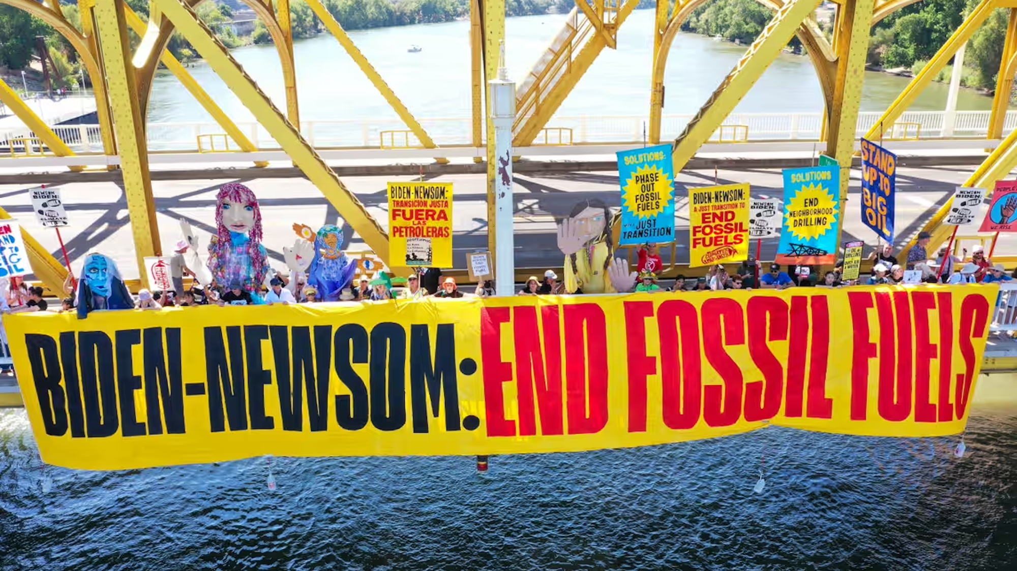 People protesting on a yellow bridge with a body of water underneath, holding a banner that says, "Biden-Newsom: End Fossil Fuels"