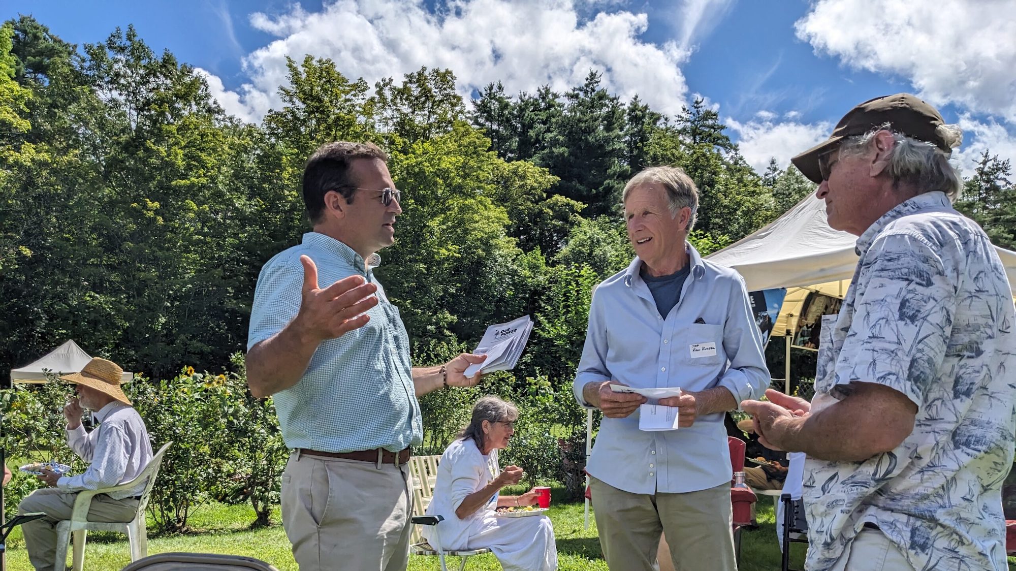 Three white men engaged in conversation on a green area with some chairs and a tent set out.