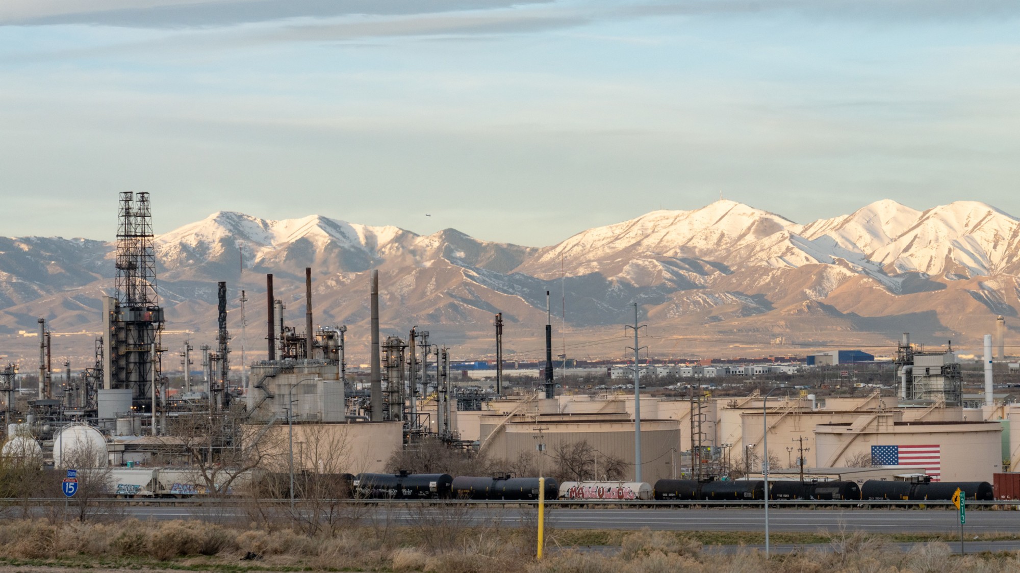 White-capped mountains tower above an oil refinery that sports an American flag.