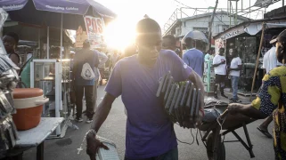 A Black man wearing a purple shirt walks through a busy marketplace holding black keyboards.