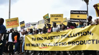 A group of protesters march with a large yellow banner that says "To end plastic pollution, we must cut plastic production."