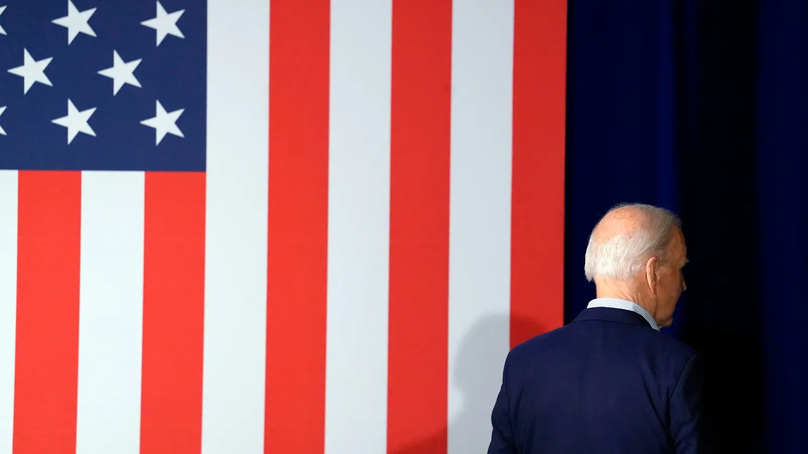 A white, balding man stands in front of an US flag.