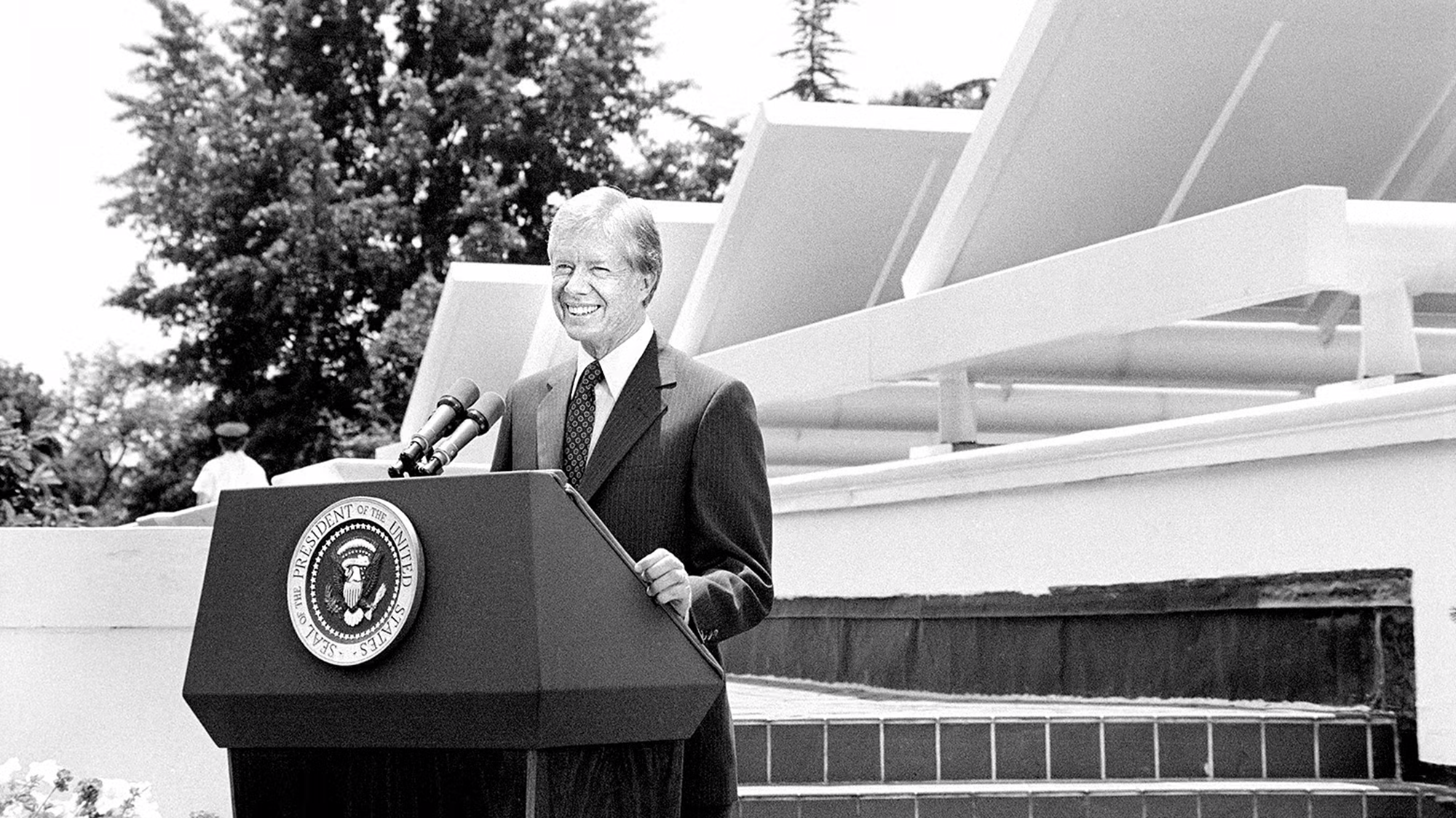 Black and white photo of man in suit in front of lectern in front of solar panels.