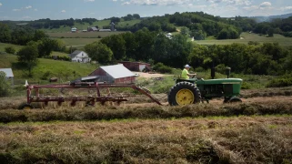 A green tractor drives through a field of yellow hay.