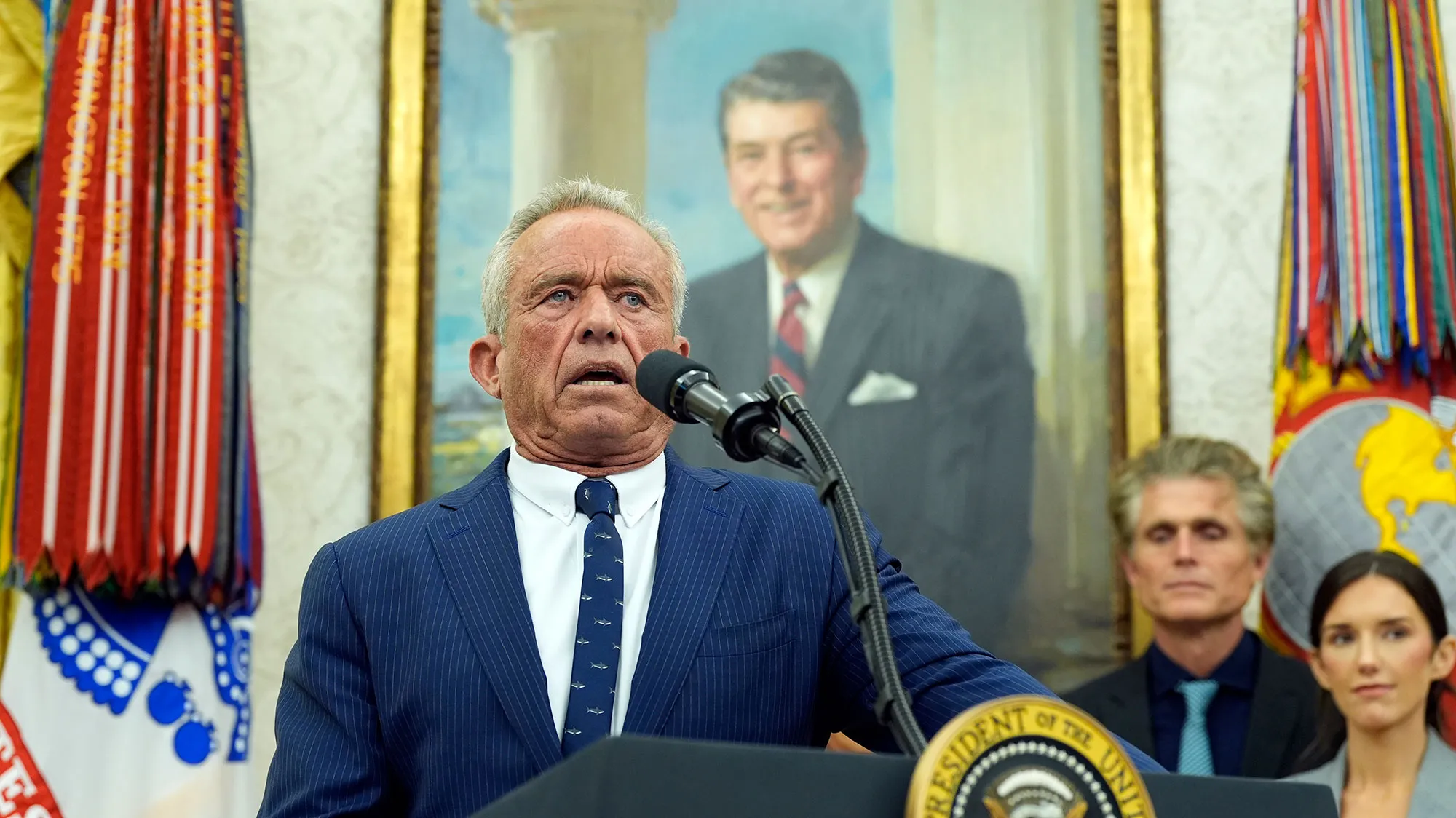 Photo of a white man standing in front of a photo of another white man.