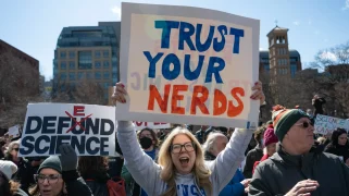 A blond person with long hair and glasses holds a sign that says "Trust your nerds"