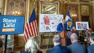 A man speaks in front of a map covered in red with a sign "Stand for our Land"