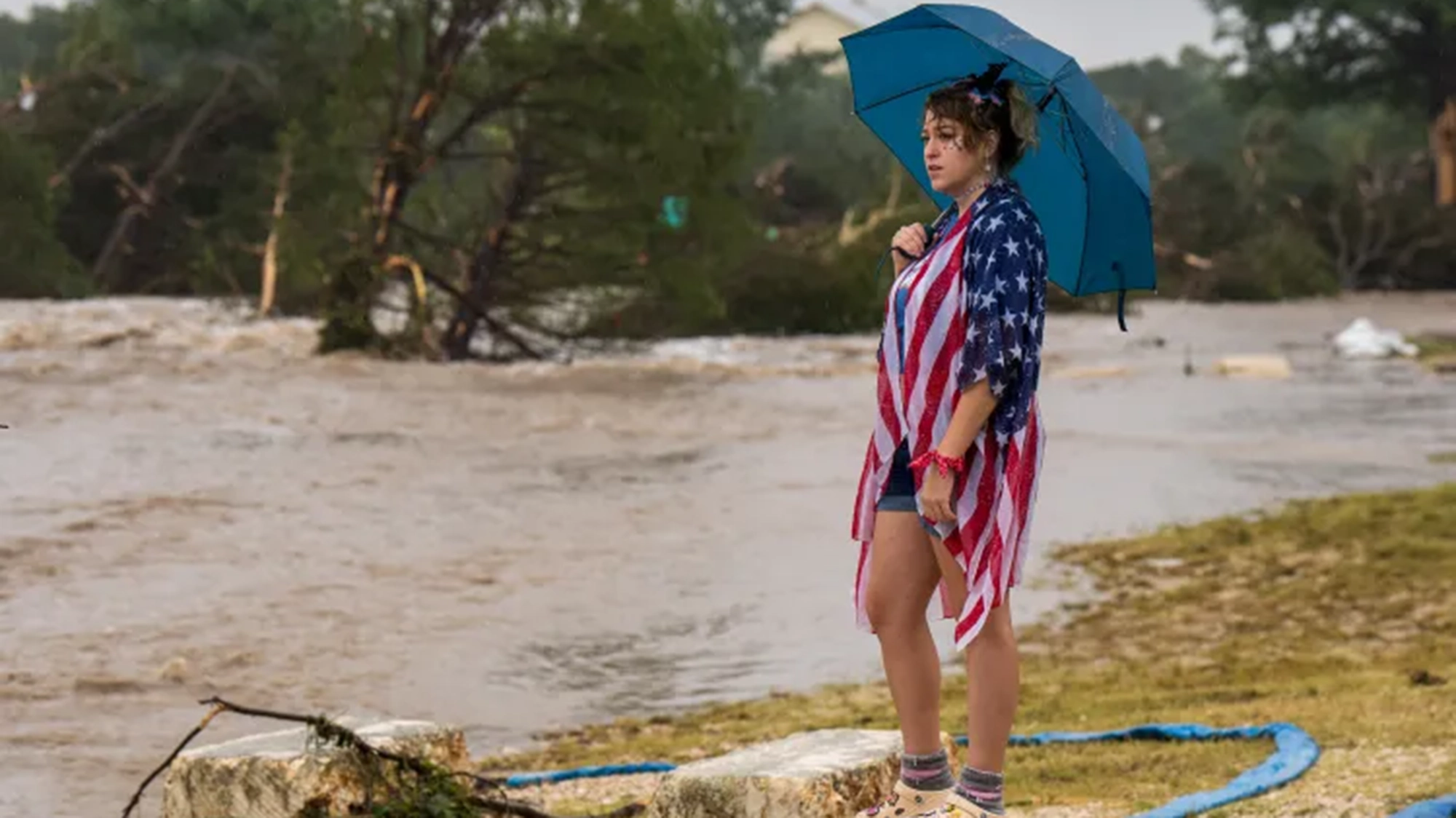 A person wearing an American flag jacket stands next to a river with an umbrella.