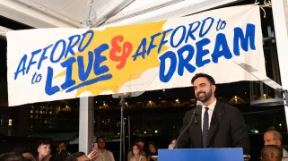 Zohran Mamdani stands in front of a banner that says "Afford to live and afford to dream"