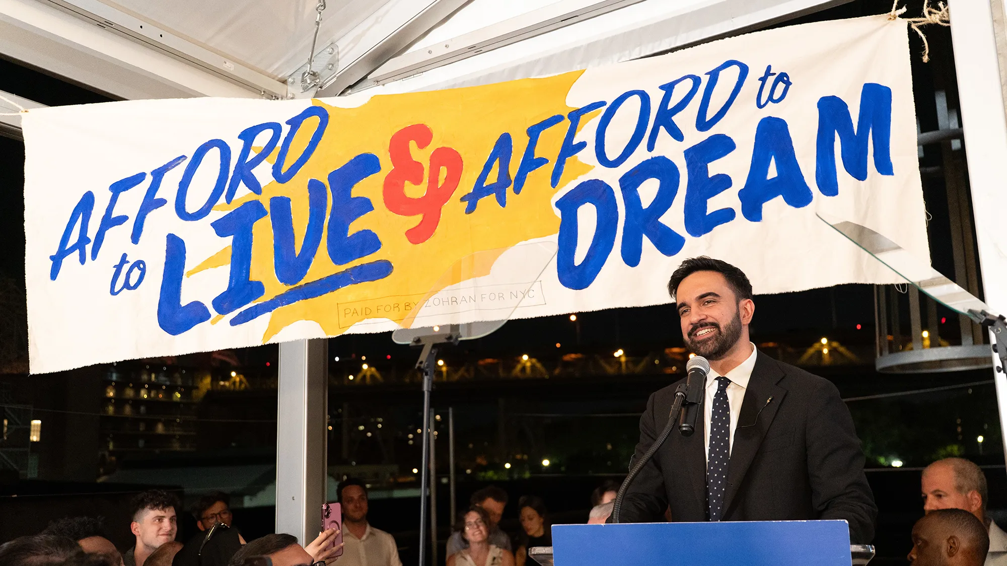 Zohran Mamdani stands in front of a banner that says "Afford to live and afford to dream"