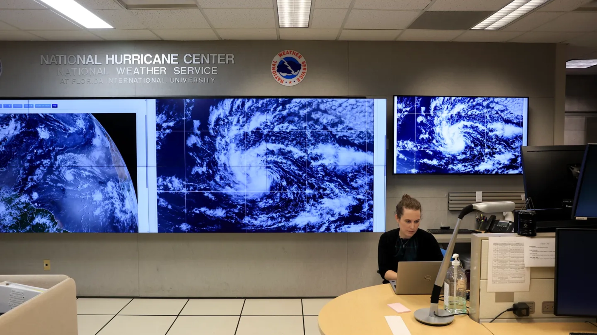 A person sits in front of a laptop in front of screens displaying a hurricane.
