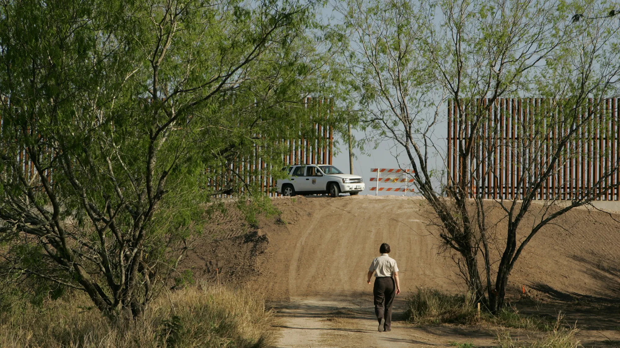 Someone walks on a dirt road towards the border wall