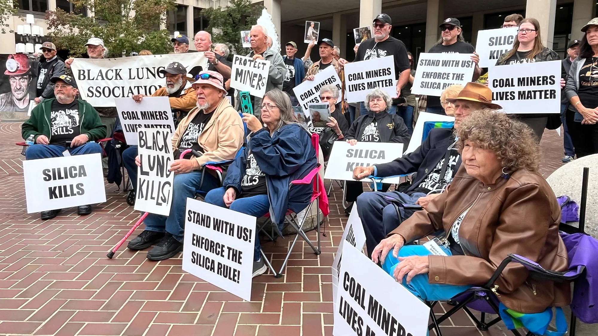 A group of people sitting and standing holding signs to raise awareness for black lung.
