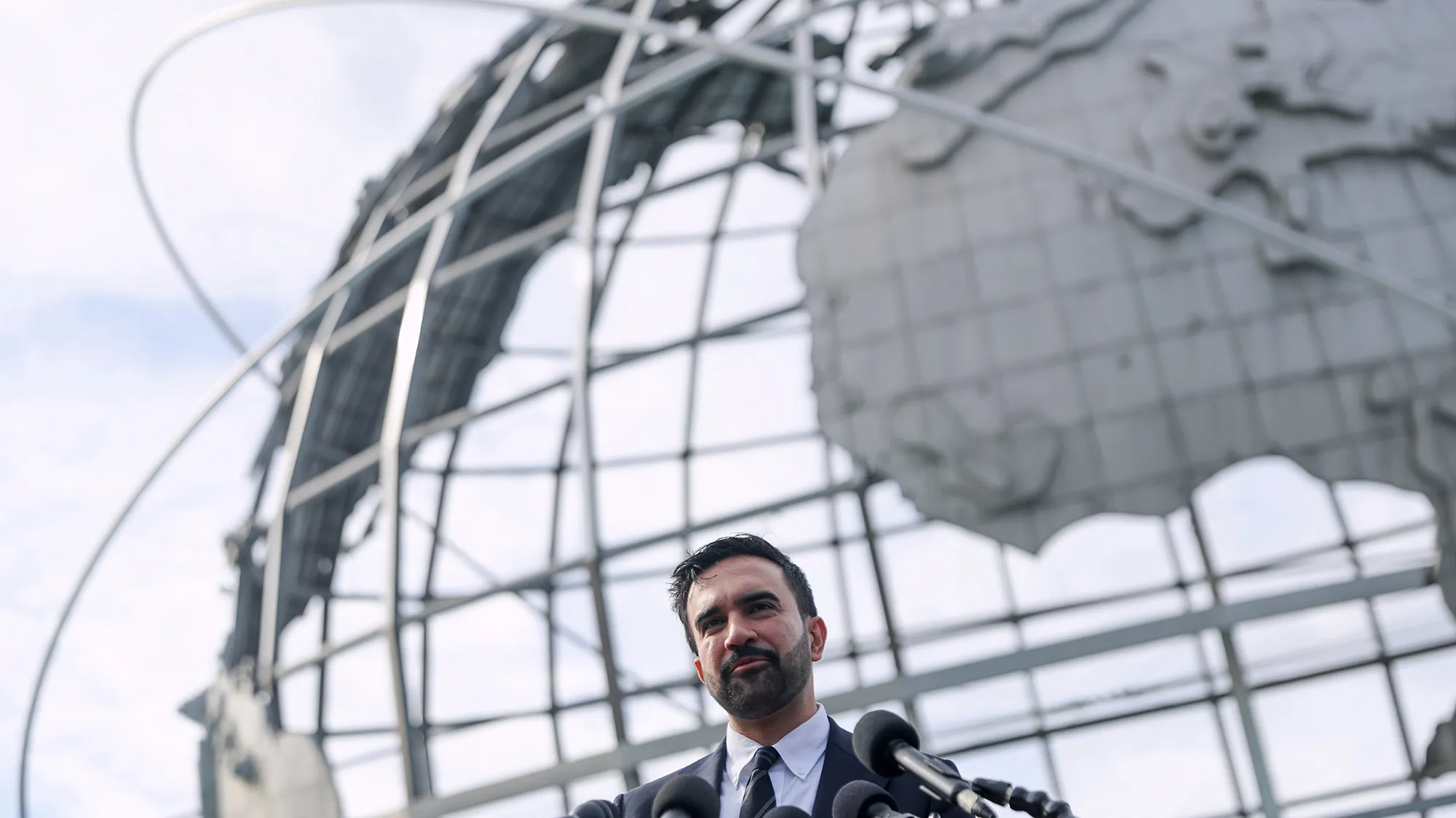 Zohran Mamdani, wearing a black suit and tie, speaks in front of a metal globe.