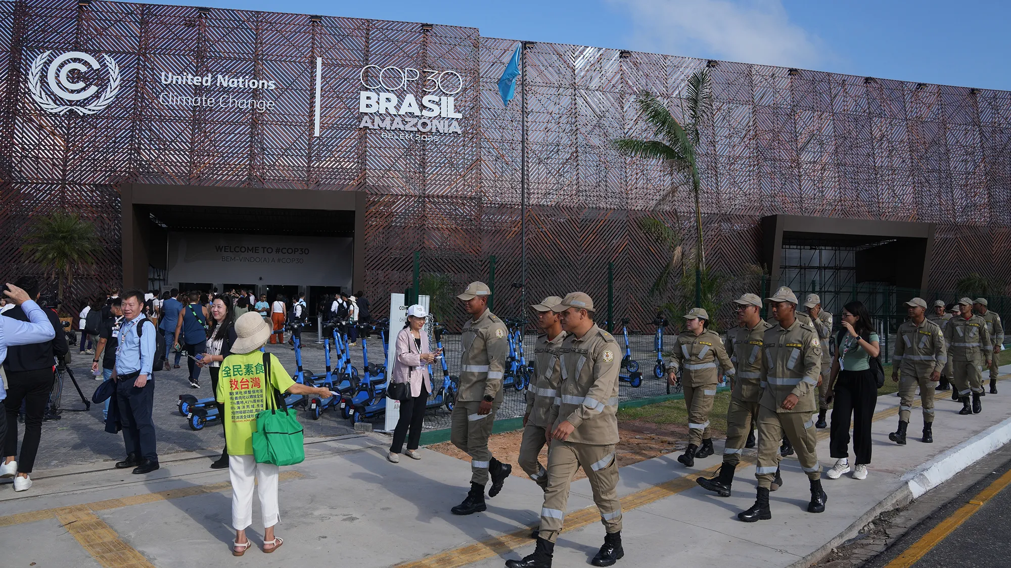 A group of people in grey-green uniforms walk past a building with a sign that says alt="A group of people in grey-green uniforms walk past a building with a sign that says "COP30 BRASIL AMAZONIA"