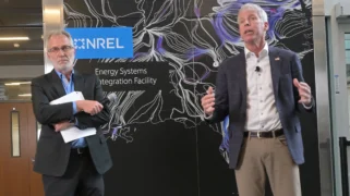 Two white men stand next to each other wearing suits. One is speaking and the other is frowning.