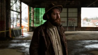 A man wearing a brown shirt and a wide-brimmed hat looks seriously into the distance. He stands in an empty warehouse.