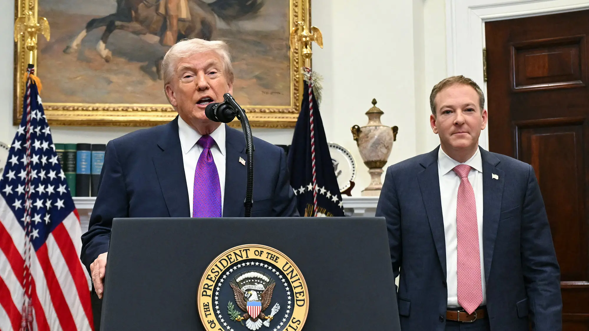 A photo of an man with white hair and tanned skin, President Trump, speaking at a podium. Next to him stands another man