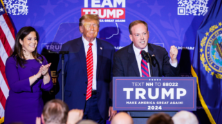 Three people stand in front of a podium that says "Trump." One, a pale white man wearing a suit, is speaking into the mic. Next to him an elderly, tan man closes his eyes. And next to him a woman, wearing a purple dress, claps.